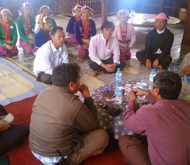 In Myanmar, a group of people sit on colourful rugs on the floor, talking in a circle around a small table