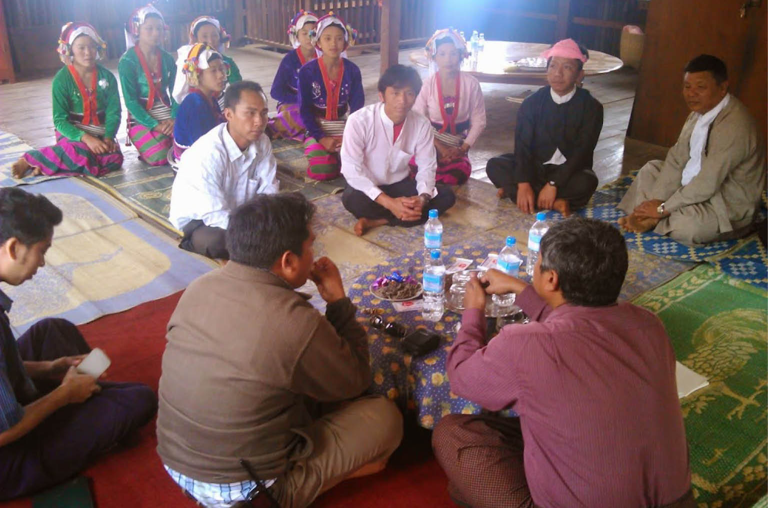 In Myanmar, a group of people sit on colourful rugs on the floor, talking in a circle around a small table