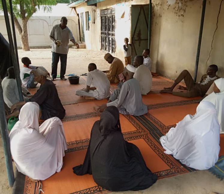 A man speaks to a group of men and women in Nigeria, gesturing openly while the group listen